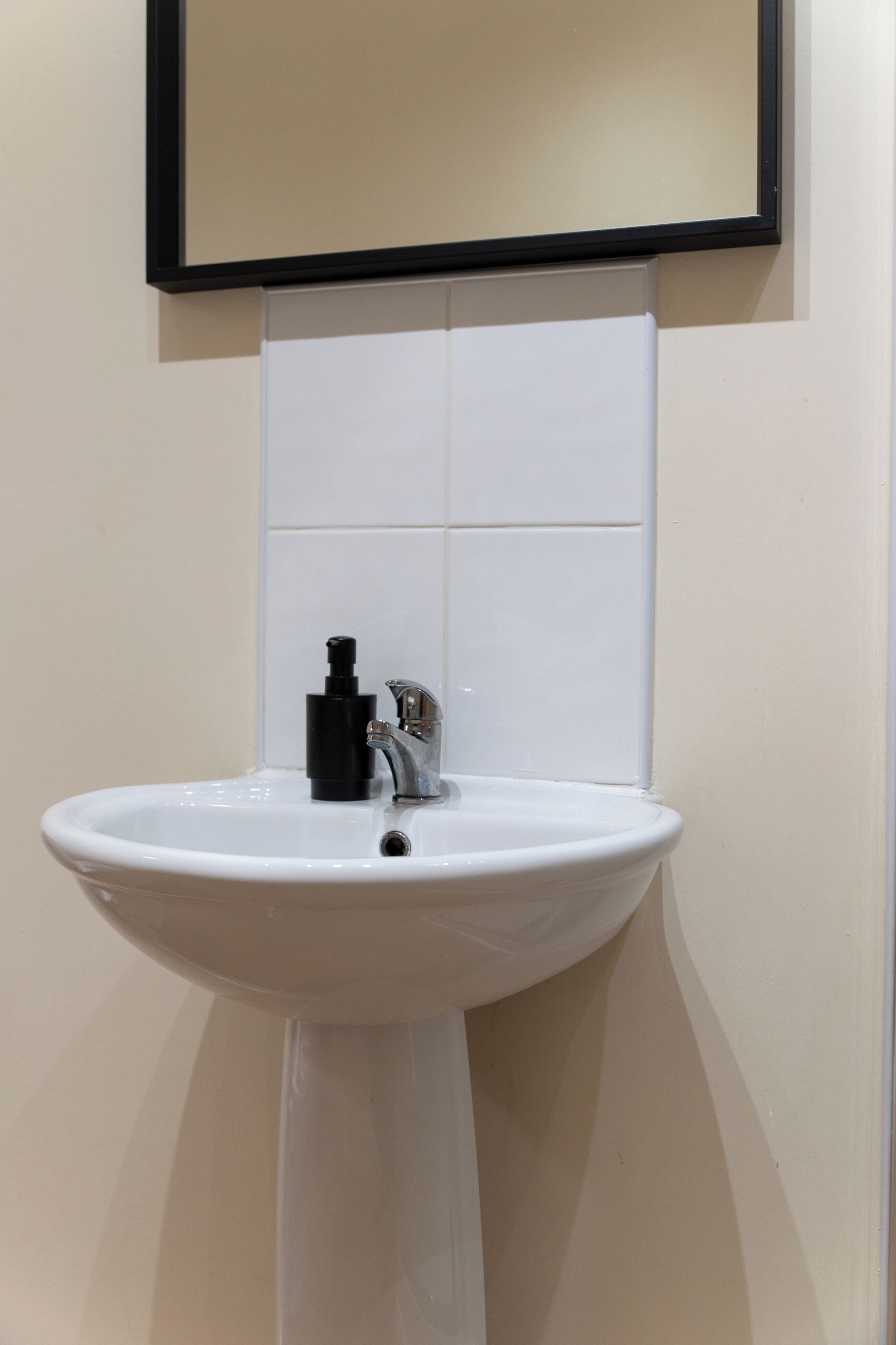 Un lavabo bianco su piedistallo con un erogatore di sapone nero e un rubinetto, montato su una parete beige rivestita di piastrelle bianche.