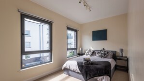 Une chambre à coucher avec un grand lit, deux fenêtres, une table de chevet et une lampe fixée au mur.