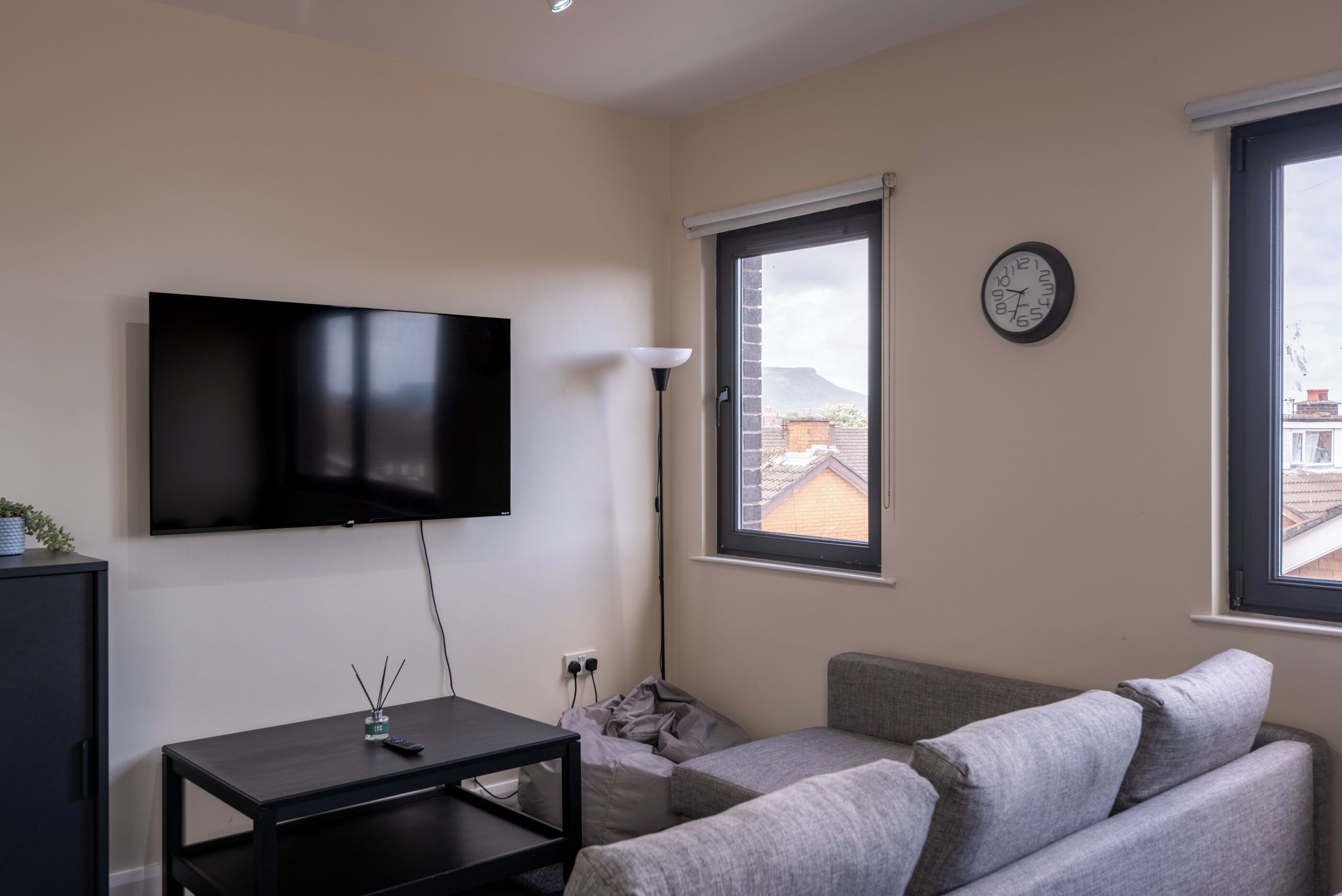 Ein Wohnzimmer mit einem an der Wand befestigten Flachbildfernseher, einer Couch, einem kleinen Tisch und einem Fenster mit Blick auf Gebäude.