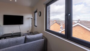 Un salon avec un canapé gris, un téléviseur à écran plat fixé au mur, une fenêtre donnant sur les maisons voisines et une horloge au mur.