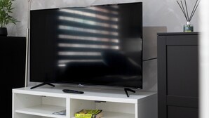Una sala de estar moderna con un televisor de pantalla plana sobre un soporte blanco, una estantería con libros y juegos de mesa, un mueble bajo y una lámpara de pie alta.