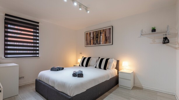 Un dormitorio con cama, mesitas de noche, una ventana con persianas, un cuadro en la pared y un estante pequeño con una planta.