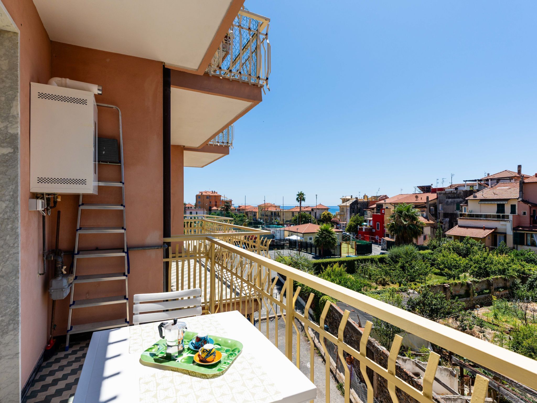 A balcony with a table set for two, a ladder, and a view of a residential area with buildings and greenery.