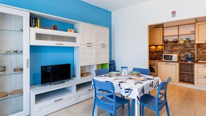 Een moderne keuken met witte kastjes, een eettafel met blauwe stoelen en een tv op een witte dressoir.