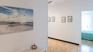 Een gang met een groot schilderij van een strand, een houten bijzettafeltje met een decoratief object, en een reeks ingekaderde foto’s aan de muur.