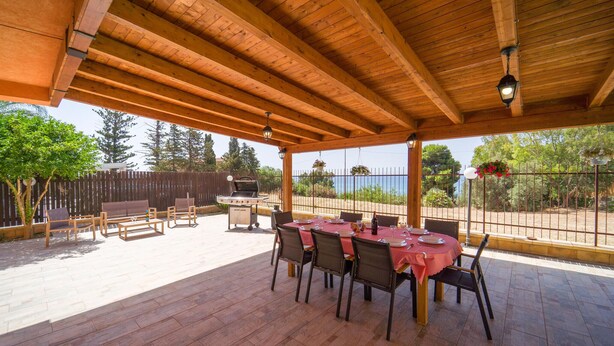Un patio exterior con techo de madera, una mesa dispuesta para comer y vista al mar.