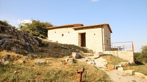 A small stone building with a tiled roof, situated on a grassy hillside with rocky terrain.