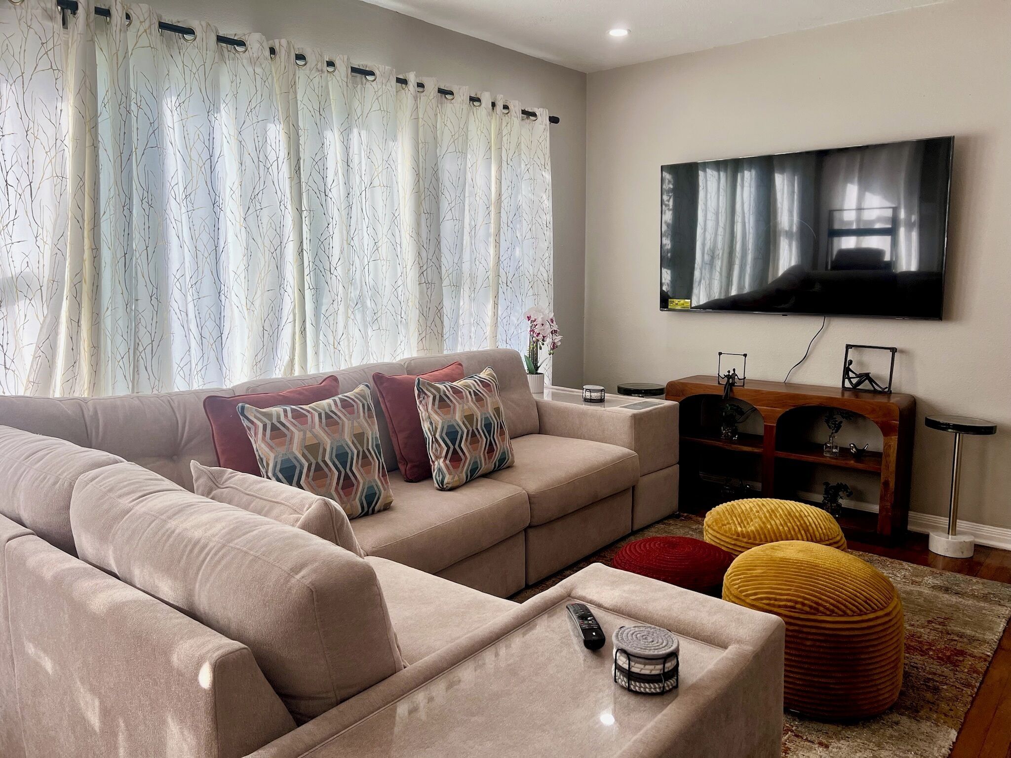 Ein Wohnzimmer mit einem beigefarbenen Sofa, gemusterten Kissen, einem Flachbildfernseher, einem Holztisch und einem roten Pouf.