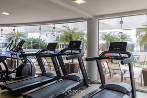 Une salle de sport équipée de tapis de course, de vélos d’exercice et d’une grande fenêtre donnant sur des palmiers.