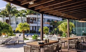 An outdoor patio with tables and chairs, surrounded by palm trees and a pool.