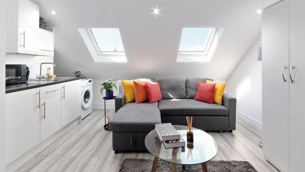 Eine moderne Wohnung mit einem grauen Ecksofa, einem runden Glastisch, einer Waschmaschine und einem Oberlicht.