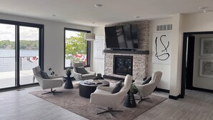 A modern living room with a fireplace, comfortable seating, and large windows overlooking a body of water. - Luxury 6BR Round Lake Home Pool, Dock, Fire Pit & Stunning Sunsets!. (Dowagiac)