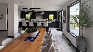 A modern kitchen with dark cabinetry, a central island, and a dining area with a wooden table and chairs. - Luxury 6BR Round Lake Home Pool, Dock, Fire Pit & Stunning Sunsets!. (Dowagiac)