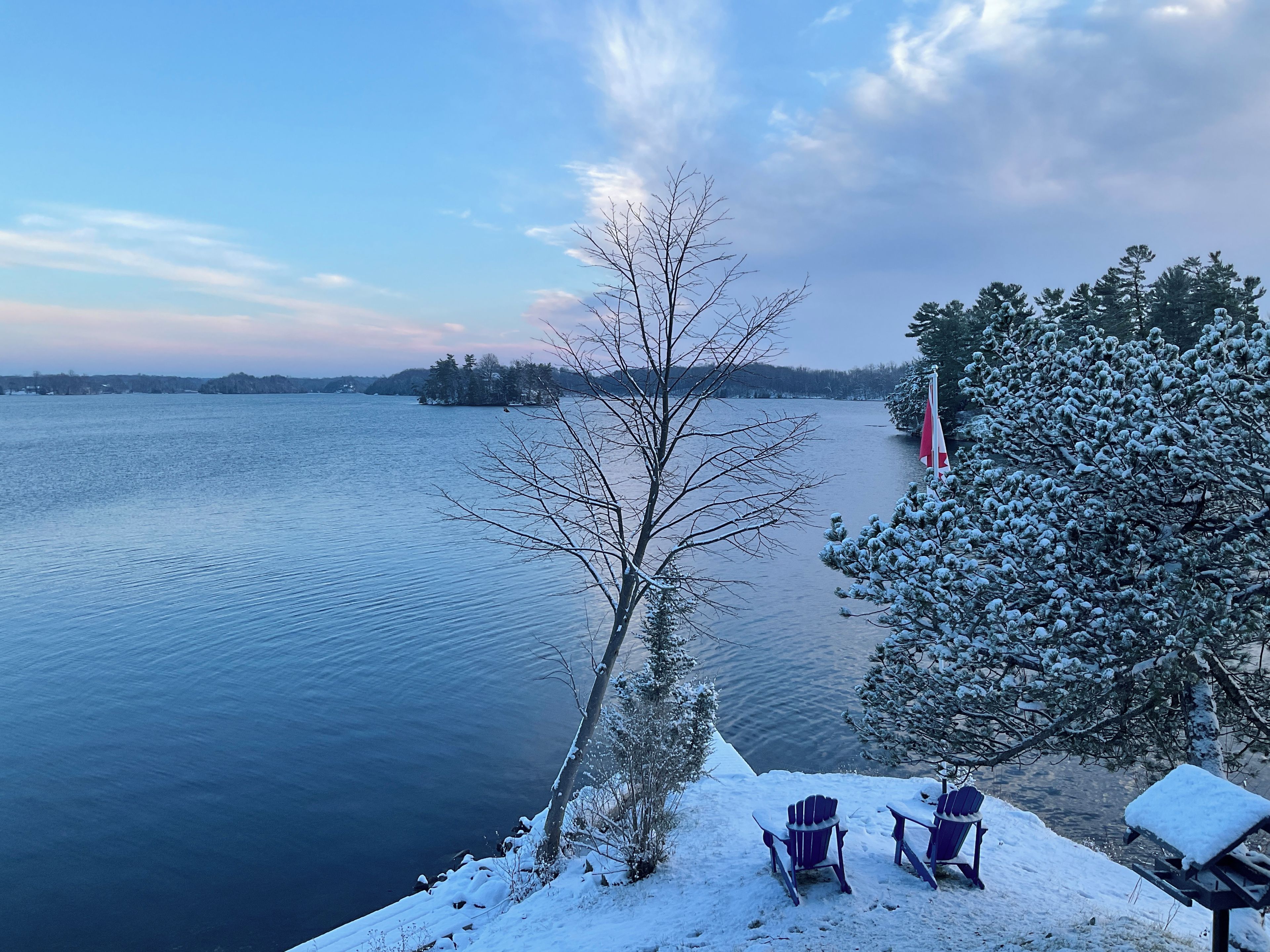 A lakeside view with snow-covered ground, two Adirondack chairs, and a flag on a dock.
