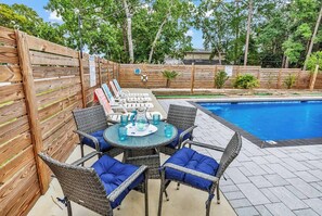 A poolside area with a round table and wicker chairs, surrounded by a wooden fence and a swimming pool. - 🌴 Stay in Style at Poolside Par | Private Pool • Mini Golf • Walk to Beach (North Myrtle Beach)