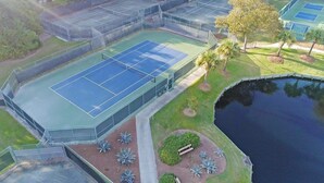 Una cancha de tenis al aire libre con una valla alrededor, un pequeño estanque y zonas ajardinadas con plantas y una banca.