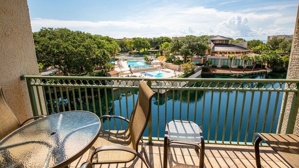 A balcony with a view of a swimming pool, outdoor seating, and a building in the background. - Spacious 2BR Condo | Balcony + Resort Amenities (Hilton Head Island)