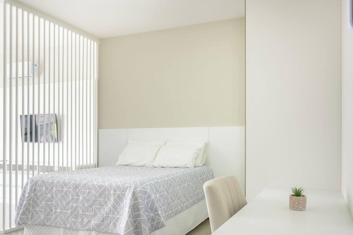 A bedroom with a bed, a small table, a chair, a potted plant, and a window with blinds.
