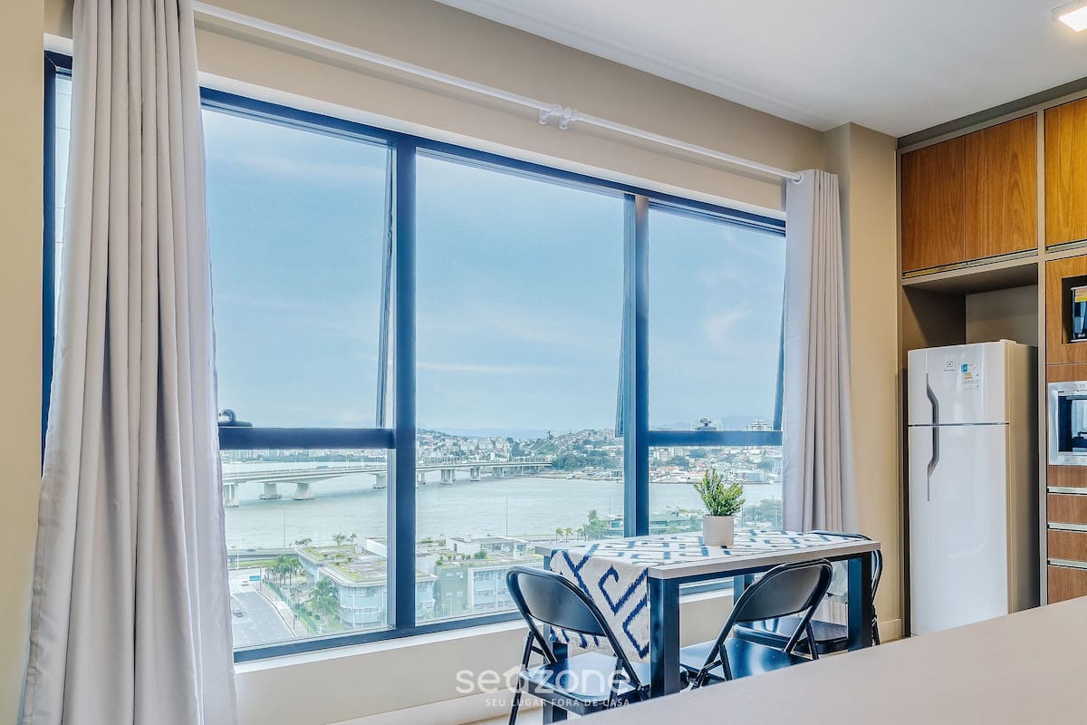 A room with a large window, a table with chairs, a refrigerator, and wooden cabinets.