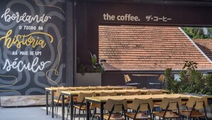 An outdoor seating area with wooden tables and chairs, a wall with text and a patterned design, and a view of a tiled roof through a window. - TVT - Luxury Studios in the Center of Florianopolis (Florianópolis)