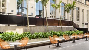An outdoor seating area with wicker chairs and tables, palm trees, and a building with large windows in the background. - TVT - Luxury Studios in the Center of Florianopolis (Florianópolis)