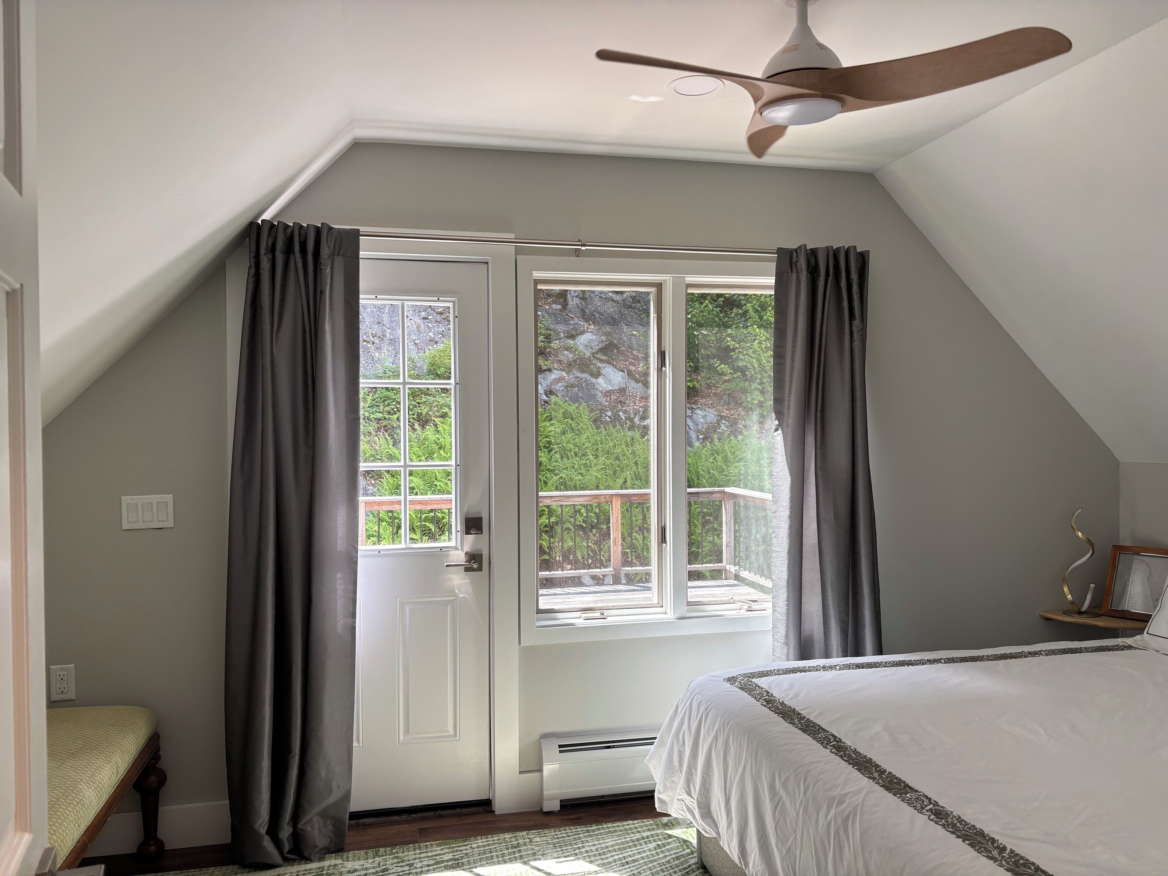 Camera da letto con un letto grande, una ventola a soffitto, una finestra con tende e vista su del verde all'esterno.