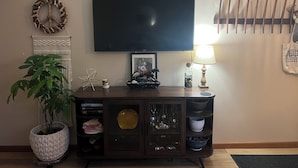 A living room with a TV, a lamp, a plant, a decorative wall piece, and a cabinet with various items. - Cozy Vibe, Clean, Beach Access, 10 mins to GOTL! (Ashtabula)