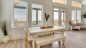 Un salón amplio con una mesa blanca, bancos y un sofá. Hay ventanas grandes con persianas, una planta en maceta y una puerta que da a otra habitación.