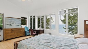 A bedroom with a large bed, a desk, a chair, a dresser, and a painting. There are multiple windows providing a view of the outdoors. - Cottage with Private Lake Access! Short distance to town (West Olive)