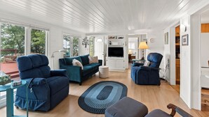 A modern living room with a teal sofa, a blue armchair, and a round ottoman. A flat-screen TV is mounted on a white cabinet, and there is a lamp on a side table. - Cottage with Private Lake Access! Short distance to town (West Olive)