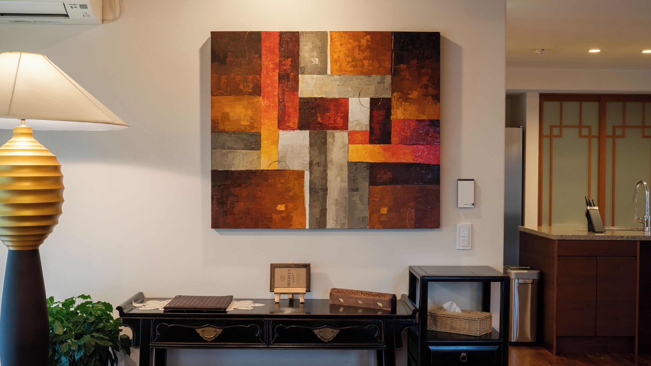 Un salon moderne avec un grand tableau abstrait, une console en bois foncé, une lampe et un coin cuisine équipé d’un évier et de placards.