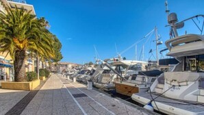 A marina with boats docked along a paved walkway, palm trees, and buildings in the background. - Blue Lagoon–4 peopl–Piscine-Tennis (Fréjus)
