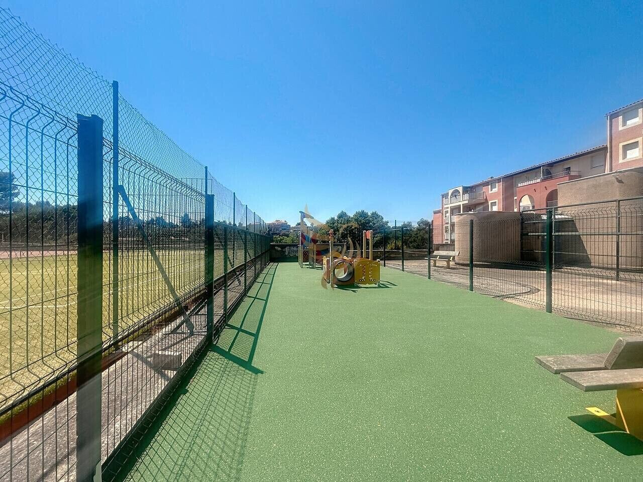 A fenced outdoor area with a green surface, possibly a playground or sports court, and a building with a red facade in the background.