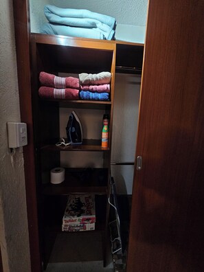 Et lille opbevaringsrum med hylder, der indeholder håndklæder, en dåse spray, et strygejern og en pakke toiletpapir.