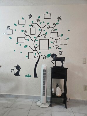 Eine Wand mit Wandbildern: einem Baum, einer schwarzen Katze und einem schwarzen Hund. Ein weißer Klimaanlage und ein schwarzer Tisch mit einer weißen Flasche darauf.