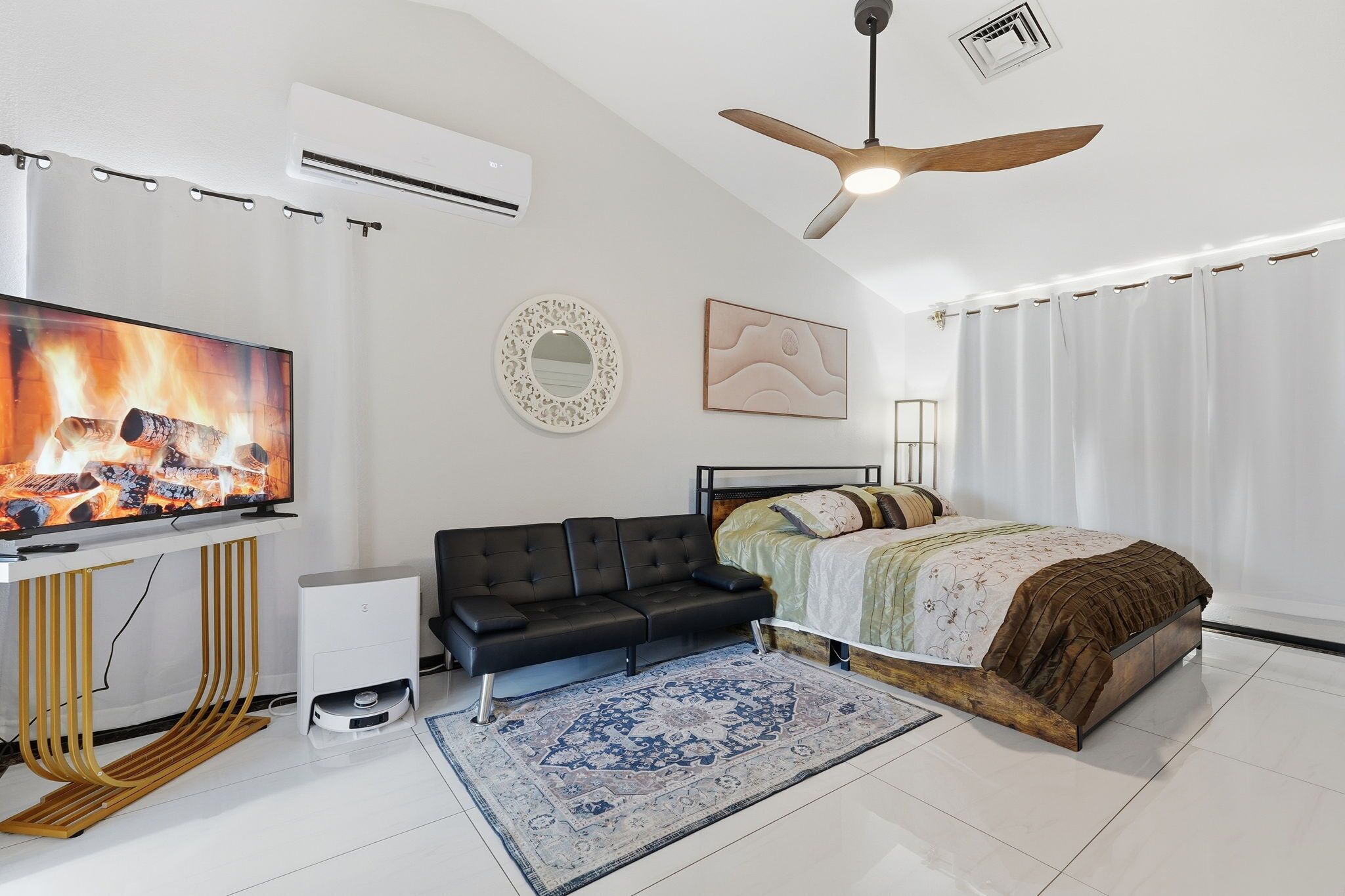 A modern bedroom with a flat-screen TV, a black sofa, a bed with a brown blanket, a patterned rug, and a ceiling fan.
