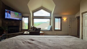 A bedroom with a large bed, a fireplace, a television displaying a weather forecast, and a view of a green landscape through a skylight. - Spacious 4-bedroom house with private Sauna, Hot tub, Fitness room in Big Sky (Big Sky)
