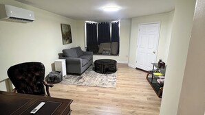 A living room with a sofa, a chair, a coffee table, and a rug. There is a door and a window with curtains.