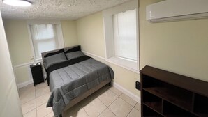 A bedroom with a bed, two windows with blinds, a ceiling light, and a wall-mounted air conditioner.