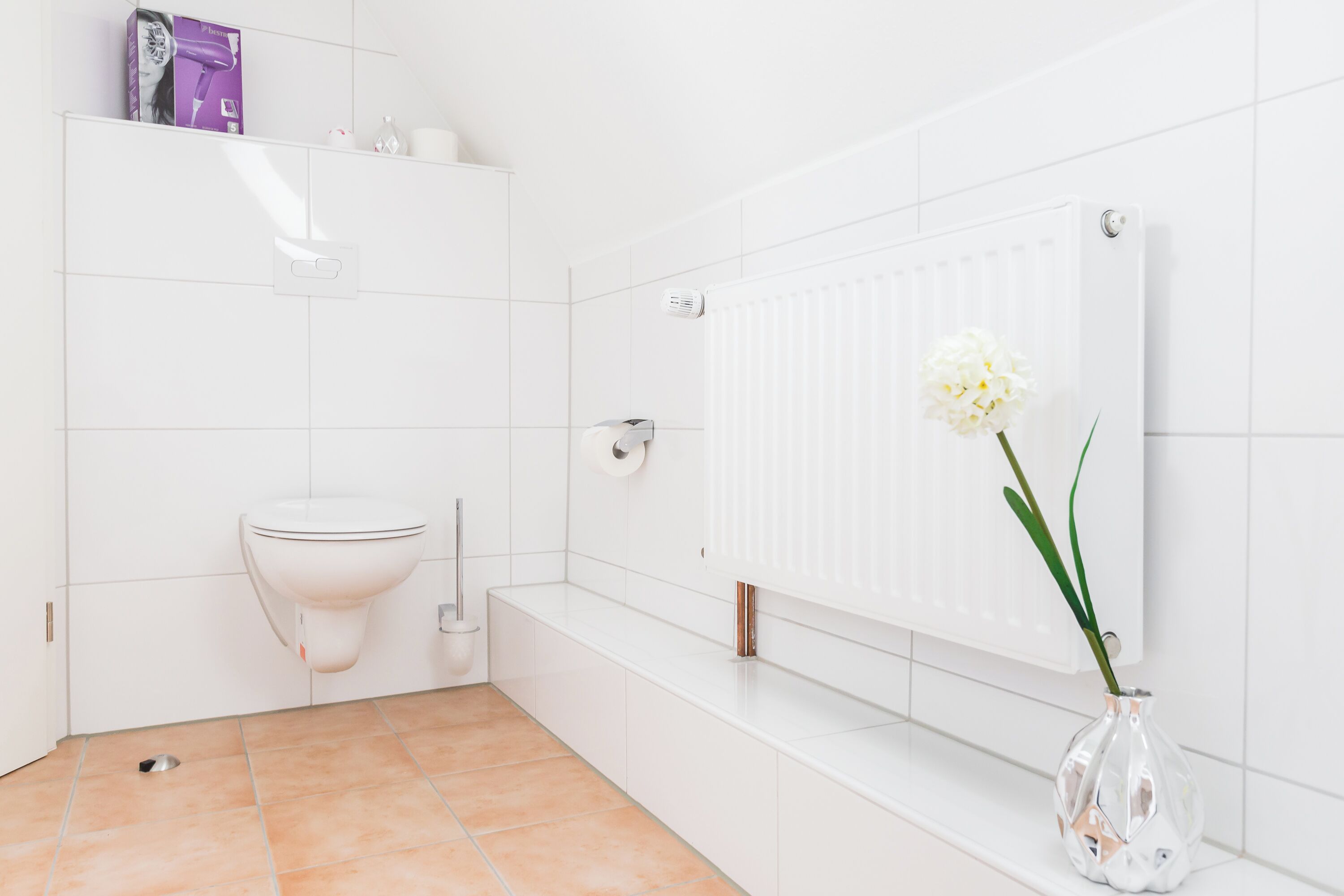 A modern bathroom with white tiles, a toilet, a radiator, and a vase with flowers.