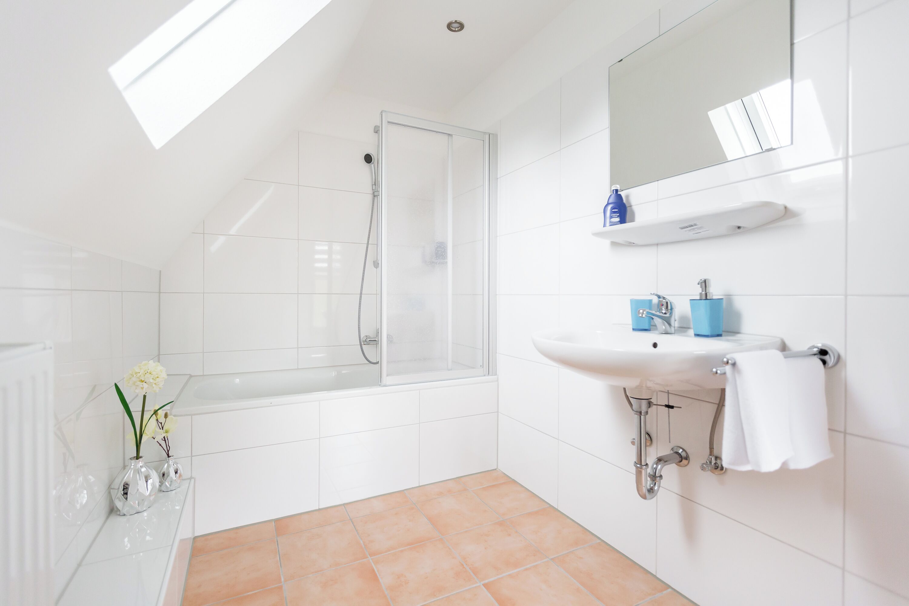 Ein modernes Badezimmer mit einer gläsernen Duschkabine, weißen Fliesen, einem weißen Waschbecken mit blauem Seifenspender und einem weißen Handtuch an der Wand.