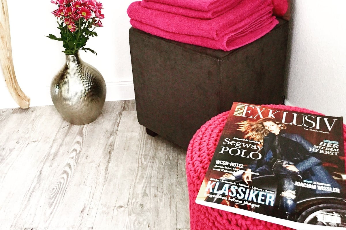 A room with a wooden floor, a dark brown ottoman with pink towels, a vase with pink flowers, and a magazine on the ottoman.