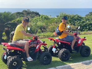 Two people on red ATVs with a scenic view of the ocean in the background. - Yadoya Lotus (Ishigaki)