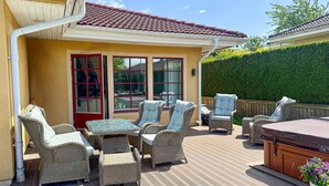 Une terrasse extérieure aménagée avec des meubles en osier, un jacuzzi et une vue sur une maison avec un toit en tuiles et une haie verte.