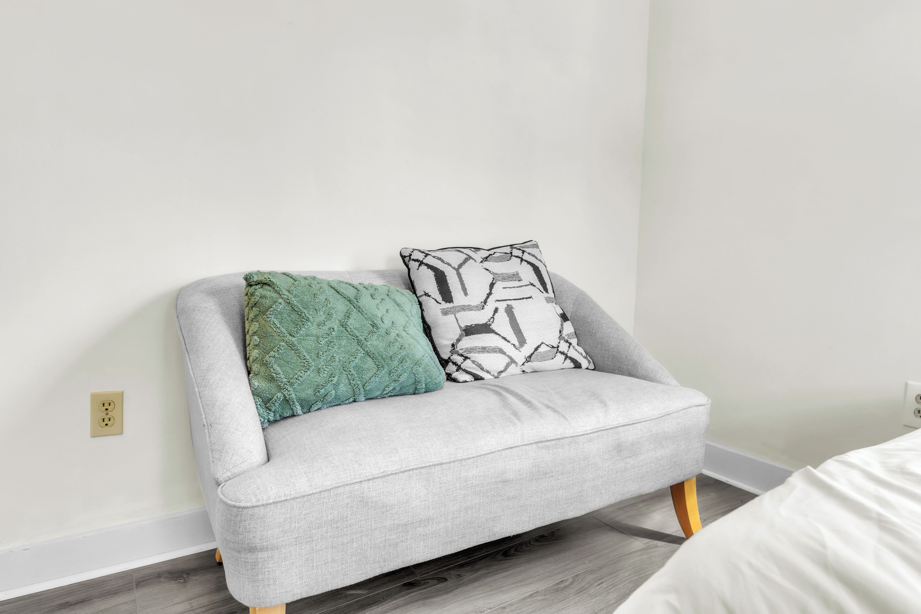 Un sofá gris con patas de madera, dos cojines estampados y una cama blanca en una habitación con una pared lisa y suelo de madera.