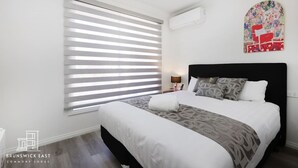 Una camera da letto con un letto grande, una finestra con persiane, un condizionatore d'aria, un comodino con una lampada e un'opera d'arte colorata appesa al muro.