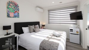 A modern hotel room with a bed, bedside tables, a flat-screen TV, an air conditioning unit, and a window with blinds.