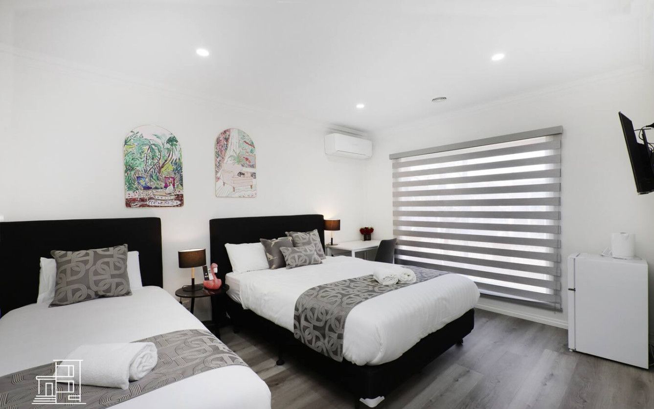 A modern hotel room with two beds, a small desk, a refrigerator, and a large window with blinds.