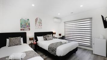 A modern hotel room with two beds, a small desk, a refrigerator, and a large window with blinds.