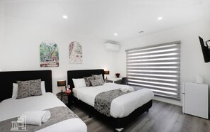 Ein modernes Hotelzimmer mit zwei Betten, einem kleinen Schreibtisch, einem Kühlschrank und einem großen Fenster mit Jalousien.